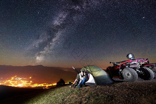 年轻的夫妇男人。女人坐着旅游帐篷前山亚视四摩托车男人。指出美丽的晚上天空完整的星星乳白色的发光的小镇背景