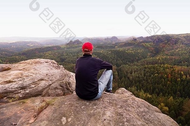 徒步旅行者男人。悬崖旅游红色的帽黑色的运动衫牛仔裤坐锋利的山悬崖谷冷早....太阳希登多云的天空
