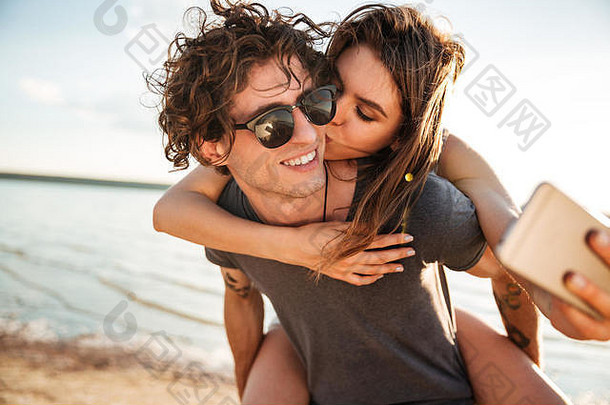 年轻的快乐夫妇爱接吻使自拍海滩肩扛骑