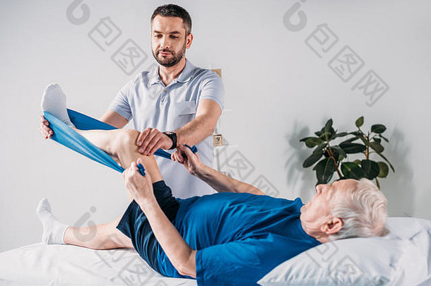 康复治疗师协助高级男人。锻炼橡胶磁带按摩表格