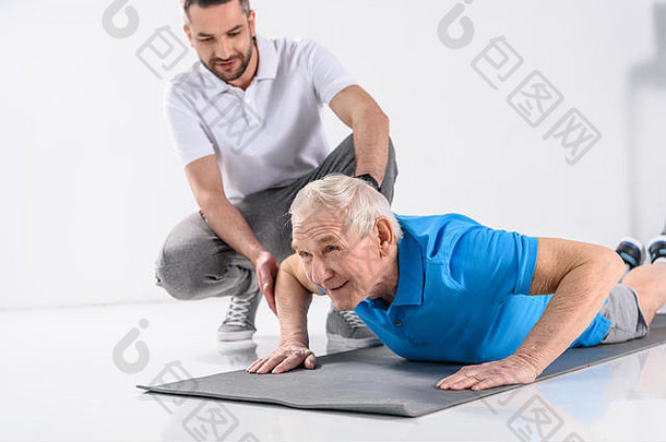 康复治疗师帮助高级男人。锻炼席灰色背景