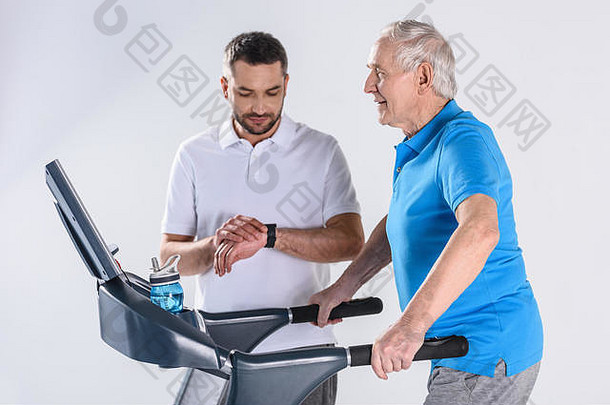 康复治疗师检查时间协助高级男人。锻炼跑步机孤立的灰色