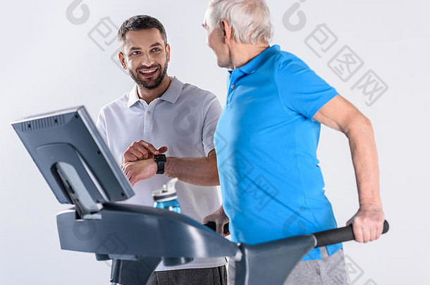 微笑康复治疗师协助高级男人。锻炼跑步机孤立的灰色
