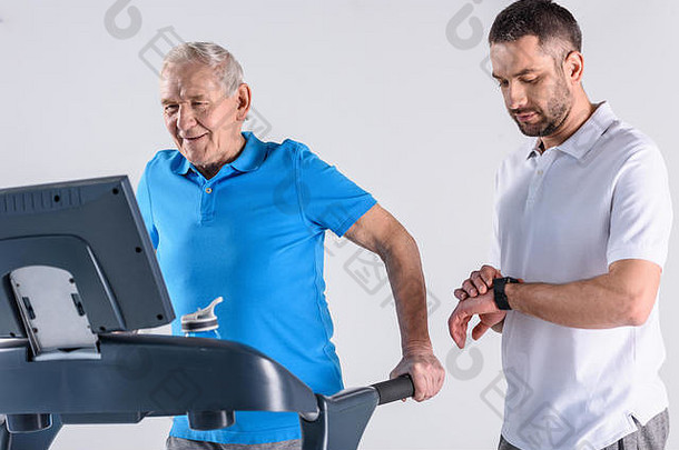 康复治疗师检查时间协助高级男人。锻炼跑步机孤立的灰色