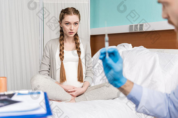 年轻的怀孕了女人坐着医院床上医生持有注射器