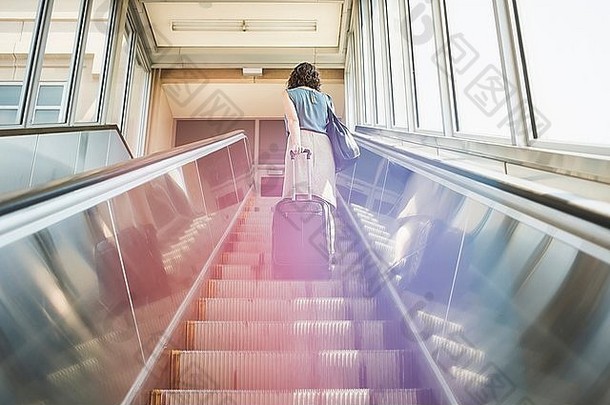 中期成人女人自动扶梯持有有轮子的手提箱后视图低角视图