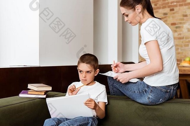 妈妈。坐着沉思的孩子持有铅笔纸书沙发上