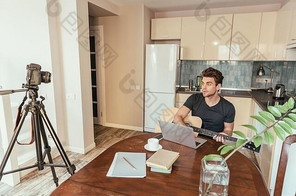 英俊的年轻的视频博客玩吉他数字相机三脚架厨房