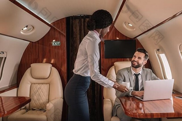 年轻的非洲美国空姐给杯咖啡微笑商人坐着移动PC飞机