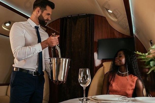 英俊的空气管家持有香槟桶瓶有吸引力的非洲美国女人私人飞机