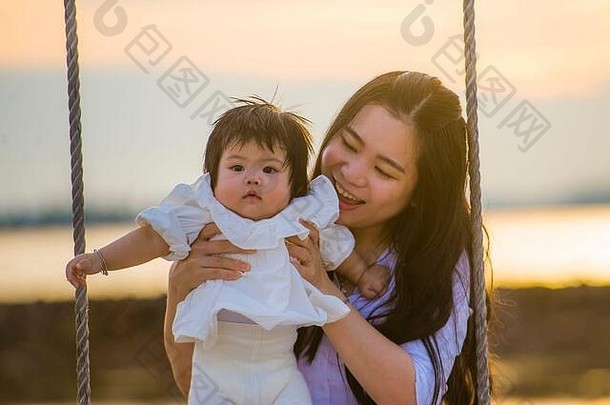 生活方式在户外肖像年轻的甜蜜的快乐亚洲中国人女人持有婴儿女孩摆动海滩摇摆不定的夏天日落妈妈。