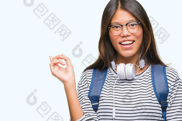 年轻的亚洲学生女人穿耳机背包孤立的背景大微笑脸指出手手指一边