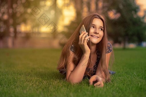 快乐电话微笑年轻的女人会说话的细胞移动电话说谎草地草草坪上城市树背景夏天晚上