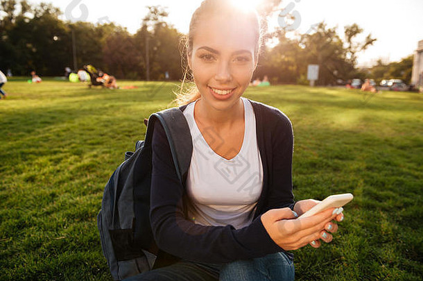 关闭肖像微笑有吸引力的学生女孩背包持有移动电话坐着草公园相机