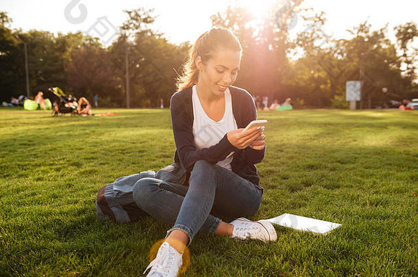 肖像快乐微笑学生女孩背包聊天移动电话坐着草公园