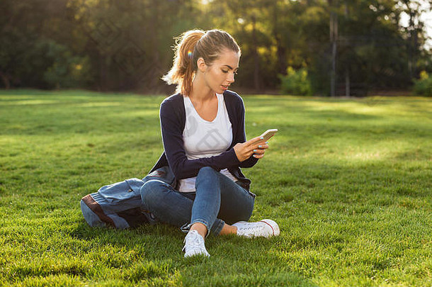肖像年轻的漂亮的学生女孩背包移动电话坐着草公园