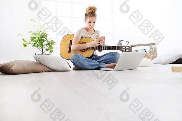 年轻的女人坐着生活房间移动电话玩<strong>吉他</strong>电脑保持首页概念
