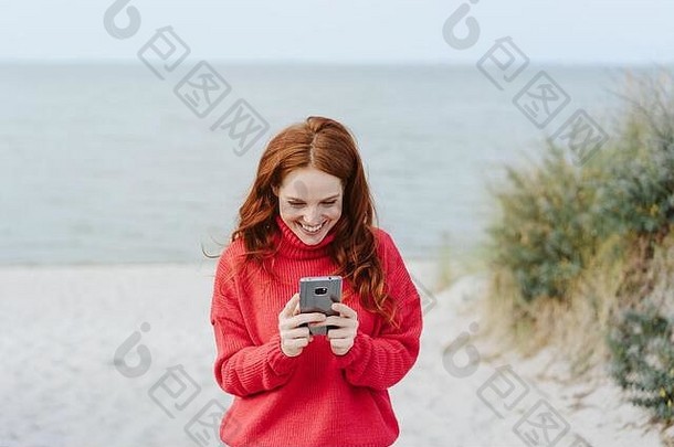 快乐年轻的女人时尚的红色的毛衣移动海边站沙丘海滩咧着嘴笑读取文本消息