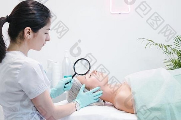 女皮肤科医生检查脸年轻的病人放大玻璃诊所美沙龙概念脸清洁剂皮肤健康