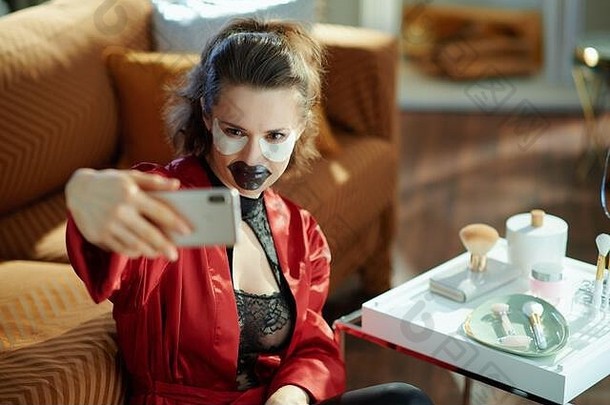 时尚的中间年龄家庭主妇黑色的身体内衣红色的浴袍黑色的水凝胶唇面具眼睛补丁采取自拍电话现代