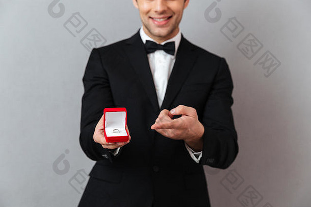 关闭肖像微笑男人。穿着燕尾服指出手指开放盒子订婚环孤立的灰色的背景