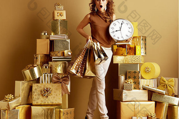 完整的长度肖像惊讶年轻的女人黄金米色裤子棕色（的）上衣购物袋时钟桩金礼物前面