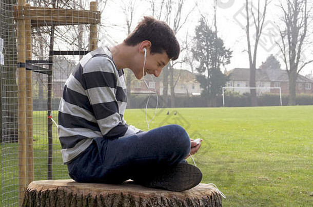 青少年男孩聪明的电话听会说话的坐着树桩英国公园少年社会媒体概念