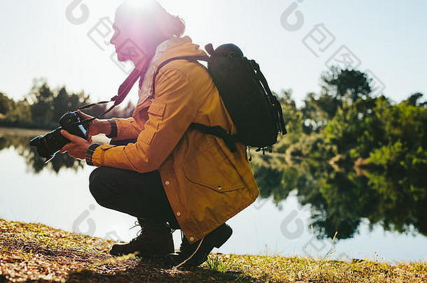 一边视图旅行者坐着湖持有数字相机男人。拍摄照片自然坐着湖阳光明媚的一天