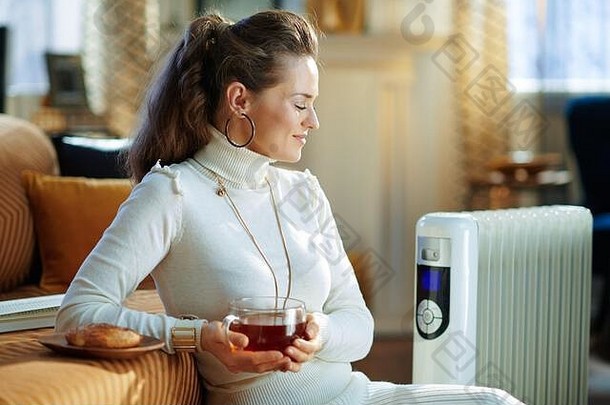 微笑时尚的中间年龄家庭主妇白色毛衣裙子坐着沙发上白色电石油散热器杯热茶好