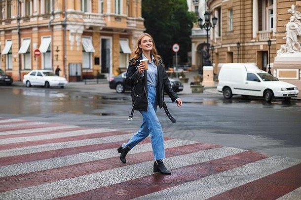 年轻的美丽的微笑女人皮革夹克牛仔裤幸福的相机持有杯咖啡手走舒适的城市