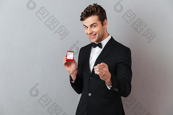 肖像满意快乐男人。穿着燕尾服持有开放盒子订婚环指出手指相机孤立的灰色的背气