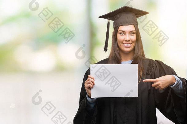 年轻的拉美裔女人穿毕业统一的持有文凭纸快乐指出手手指