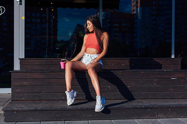 时尚的快乐年轻的浅黑肤色的女人女人穿白色短裤运动鞋持有粉红色的杯<strong>咖啡</strong>坐着<strong>咖啡</strong>商店