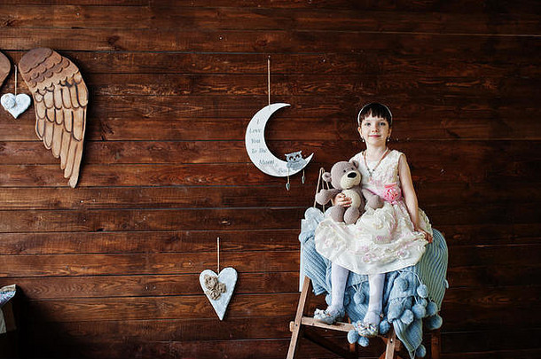 一个穿着衣服的美丽女孩的肖像，手里拿着一个玩具，坐在靠着木墙的木梯子上。