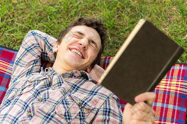年轻的男学生躺在公园里，手里拿着书，笑着