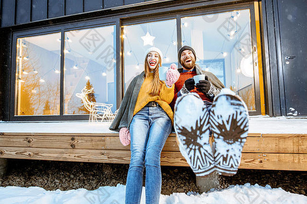 寒假期间，一对年轻夫妇坐在山上现代住宅的露台上，穿着格子布，喝着热饮热身