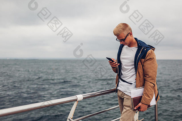 在敖德萨，一名大学生背着背包和书，用手机拍摄大海，欣赏海滩上的景色。在海滩上散步的游客