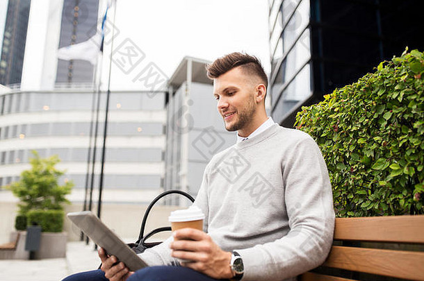 坐在城市街道长椅上的男子，手里拿着平板电脑和咖啡