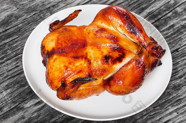 美味的金黄色酥皮整只鸡在烤箱中烤熟，放在旧的深色木制桌子上的白色盘子上，俯瞰，特写
