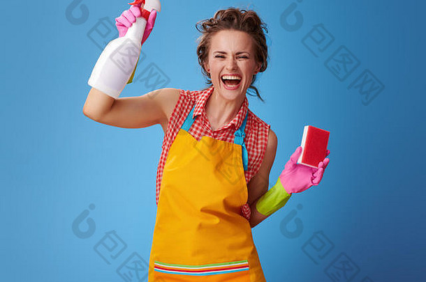 <strong>大扫除</strong>时间。身着黄色围裙、厨房用海绵和一瓶清洁剂的年轻女子微笑着，欢欣地站在蓝色的地面上