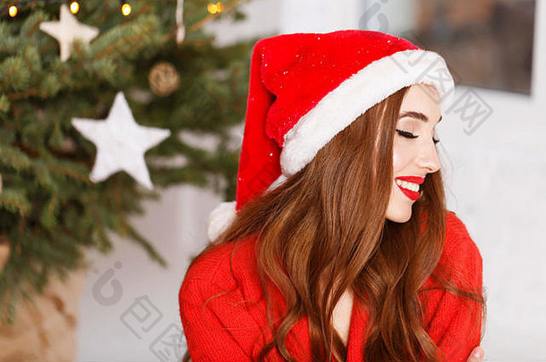 穿着圣诞帽和红色毛衣的迷人圣诞妇女的肖像坐在舒适的新年云杉旁的地板上，新年、圣诞节、假日、纪念品、礼物、<strong>购</strong>物、折扣、商店、雪人圣诞老人、化妆、发型、<strong>狂欢</strong>节。