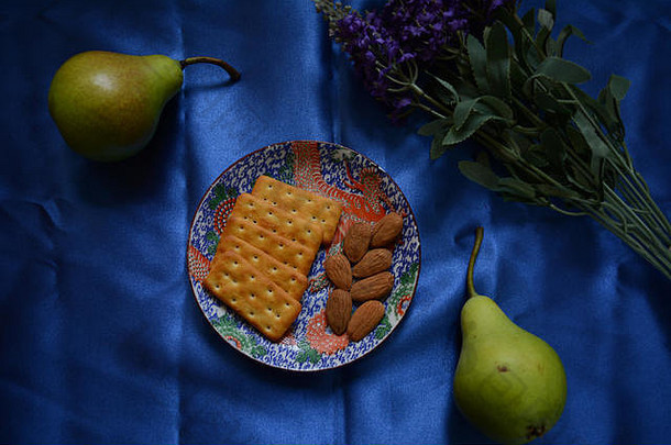静物手指食物摄影，平放。饼干、梨、杏仁和薰衣草作为装饰。蓝色背景，闪亮的丝绸布料。