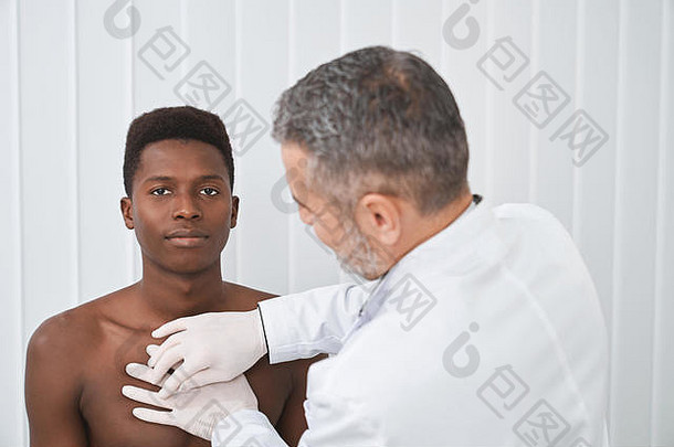 一名非洲患者在医务室看摄像机，摆姿势，坐在检查台上。身穿白色长袍、戴着手套的专业医生，对男子胸部进行触诊和诊断。