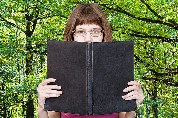 戴眼镜的女孩读着一本大书，书的封面是空白的，背景是绿色的夏日森林
