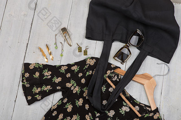 时尚潮流-太阳镜、黑色环保手提包和衣架上印有花卉图案的连衣裙、珠宝：发夹、白色木制桌上的银和金耳环