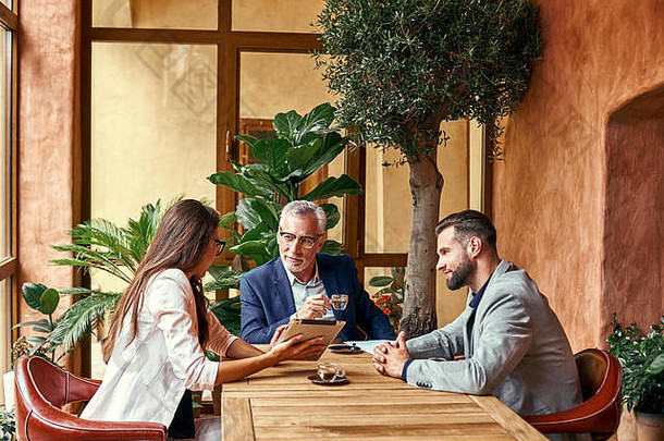 商务午餐。餐厅里的三个人坐在桌旁喝着热咖啡，一位女士在数字平板电脑上展示创意。团队合作理念