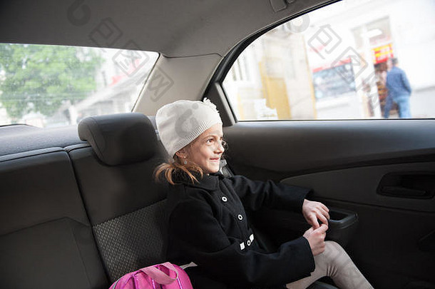 快乐的小女孩，穿着秋天的黑色外套，戴着白色帽子，坐在安全车的后座上，背着粉红色的背包，微笑着离开学校