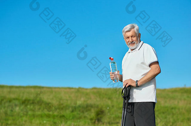 那个老人拿着水瓶和跟踪棒。站在背景上绿色的草地上。穿着带深蓝色条纹的白色马球衫、黑色长裤、运动鞋。看着摄像机。