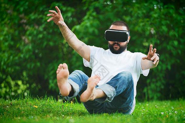 戴着虚拟现实护目镜、留着胡子的年轻人坐在花园里。生活方式VR乐趣和放松概念。
