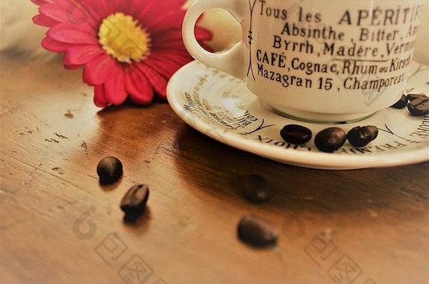 一个小的白色咖啡杯，上面有法国开胃酒，放在一张木桌上，旁边放着咖啡豆和一朵粉红色的花。